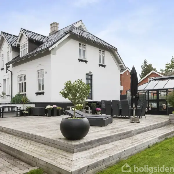 Huset har en hvid facade og charmerende sort tag, der står i en have med en stor træterrasse. Terrassen er indrettet med havemøbler og planter, omgivet af grønne områder.