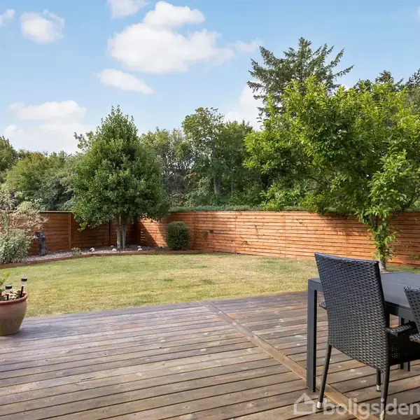 Terrassebord med sorte fletstole står på en træterrasse, der grænser op til en græsplæne omkranset af et lavt plankeværk. Træer og buske i baggrunden under blå himmel med hvide skyer.