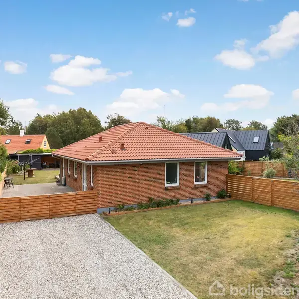 Et rødt murstenshus med tage af tegl er placeret i en have med grusindkørsel. Omgivet af hegn og træer i et boligkvarter.