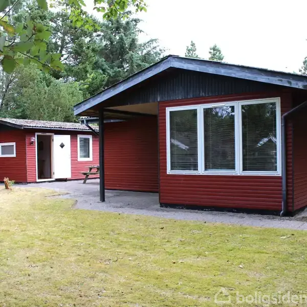 Rødt træhus står stille i en grøn have med græs og omgivet af træer, har store vinduer og en skorsten. Ingen tekst er synlig i billedet.
