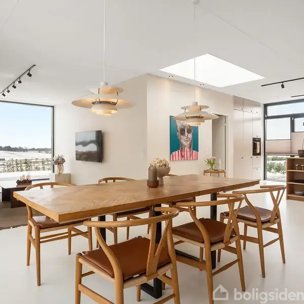 Et spisebord med seks træstole står i et lyst køkken-alrum. Over bordet hænger to moderne lamper. I baggrunden ses et køkkenområde og store vinduer med udsigt.