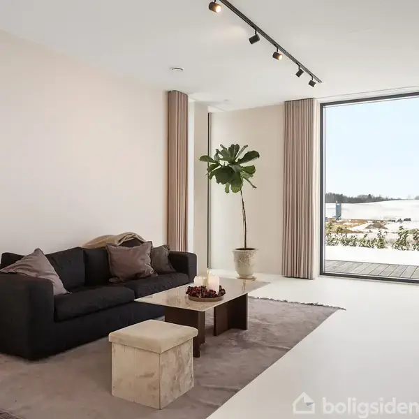 Sort sofa med puder står i stue med beige tæppe. En gulvlampe og en plante står ved siden af. Store vinduer giver udsigt til snedækket landskab.