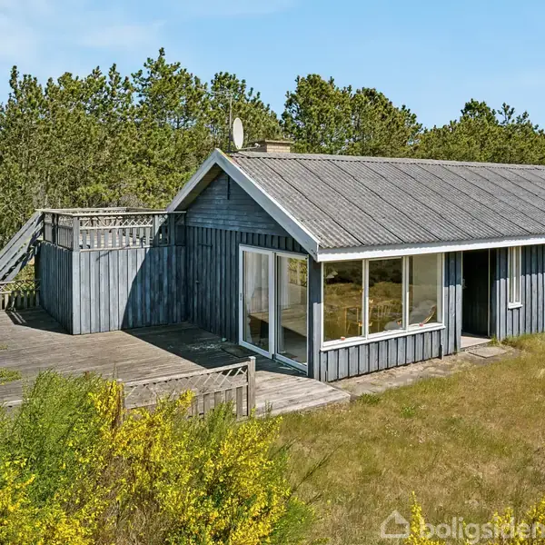 Et træhus står stille, omgivet af buske og træer. Det har et stor vindue og en træterrasse, hvor man ser skov i baggrunden.
