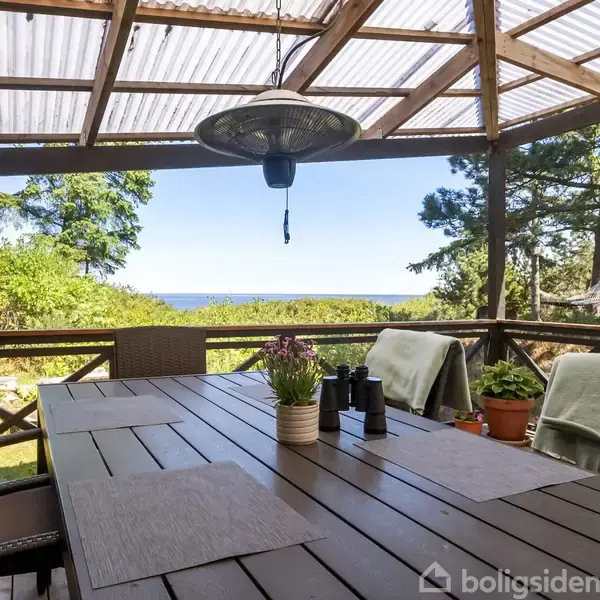 Et stort træbord står på en overdækket terrasse med havudsigt. Omkring bordet er stole med tæpper, og blomster og krydderier pynter i midten. Skov og hav ses i baggrunden.