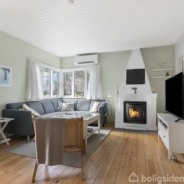 Stue med grå hjørnesofa, tændt brændeovn og stor fladskærms-tv på et tv-bord. Rummet har lyse trægulve, hvidmalet loft og vægge med naturbillede ved siden af dør.