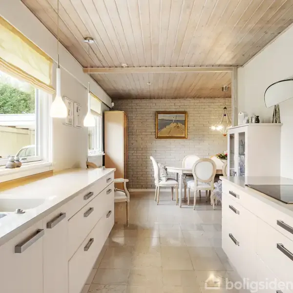 Køkken med hvide skabe og skuffer, arrangeret i en galley-stil. Et spisebord med stole står i baggrunden. Store vinduer til venstre slipper naturligt lys ind.