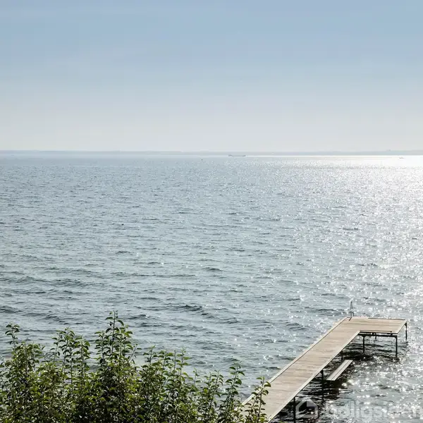 Havet glitrer i solen, skaber en rolig atmosfære. En træbro strækker sig ud i vandet til højre, omgivet af frodig vegetation.