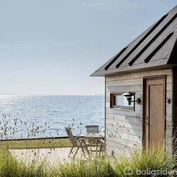 Et gråt træhus står ved havet. En lille terrasse med stole og bord ses i forgrunden, omgivet af græsset. Vandet glitrer under en klar himmel.