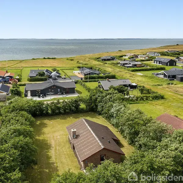 Hus set fra oven omkranset af træer, beliggende i et sommerhusområde ved kysten. Græsplæner omringer husene, der ligger tæt på havet og med udsigt til vandet.