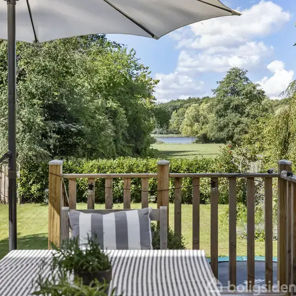 Parasol står over terrassebord med stribede hynder, placeret på en terrasse med udsigt over en have og en skovklædt sø i baggrunden.