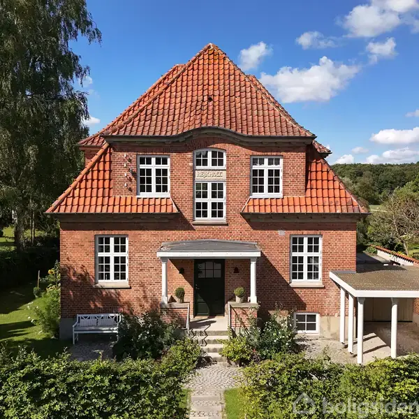 Rødt murstenshus med asymmetrisk rødt tegltag og hvide vinduer; står foran en garage og omkranset af grønne hække med en blå himmel og skov i baggrunden.