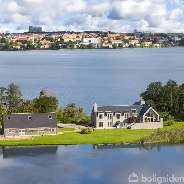 Et moderne hus ligger ved en sø med en stor have, omgivet af træer. I baggrunden ses en by med mange bygninger og skyer på himlen.