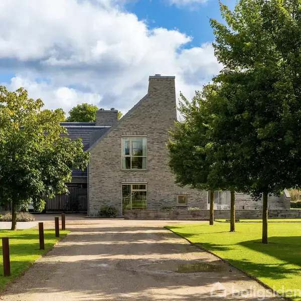 En moderne villa og et anneks ligger ved bredden af en sø, omgivet af grønne træer og græs. I baggrunden ses en by med bygninger under en blå himmel med skyer.