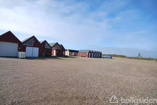 Røde lagerbygninger står i række på en grusplads under en klar blå himmel. En bil er parkeret i baggrunden nær bygningerne, som omgives af åbent, fladt terræn.