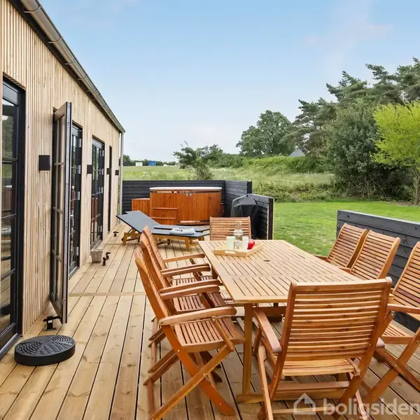 Terrasse med træmøbler, hvor en række stole og et bord står placeret. Aktive skodder åbner mod huset. Omgivet af grønt landskab og en hottub i baggrunden.