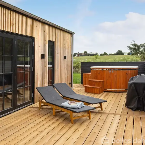To liggestole står på en træterrasse ved siden af et moderne kolonihavehus. I baggrunden ses et spabad og en gasgrill. Omkringliggende natur med græs og træer.