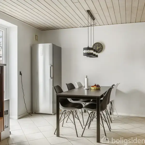 Køkken med sort spisebord og seks stole står på klinkegulv ved siden af et højt køleskab. På væggen hænger et ur, og en lampe hænger over bordet.