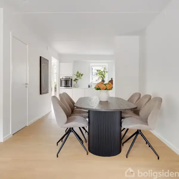Ovalt spisebord med grå stole står stille på lyst trægulv. I midten af bordet er en vase med blomster. I baggrunden ses et vindue og et billede på væggen.