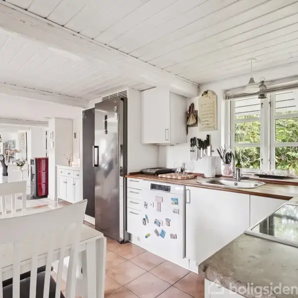 Køkken med hvide skabe og mørke bordplader, der har køleskab og komfur. Spisebord med hvide stole i forgrunden. Store vinduer giver udsigt til grønne planter udenfor.