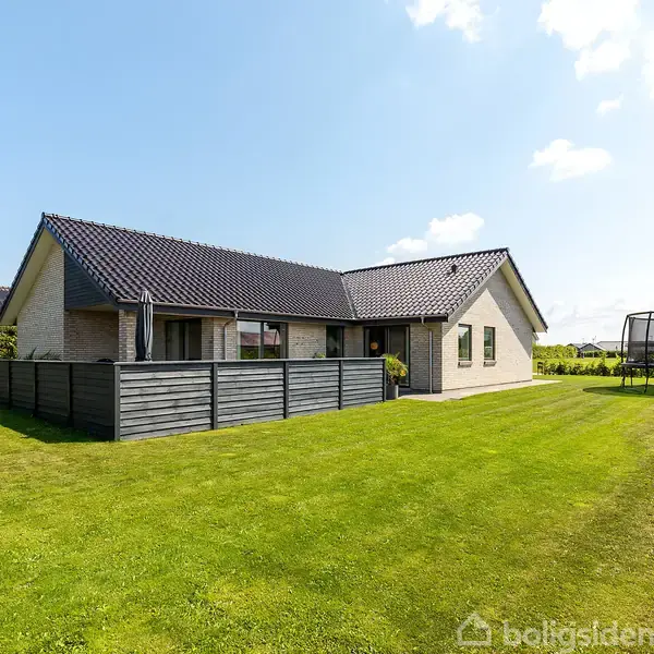 Et hus har en gråmalet facade og sort tag. Det står stille på en velplejet græsplæne omgivet af hække. En trampolin er placeret til højre for huset.