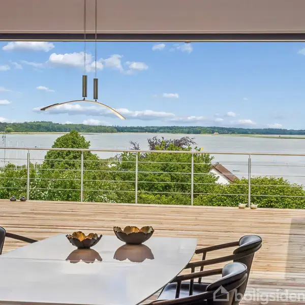 Et spisebord med stole står stille foran en stor glasvæg. Udsigt over en terrasse med gelænder, græs og vand i baggrunden under en klar blå himmel.