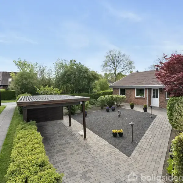 En rødstensbungalow står ved siden af en enkelt carport. Haven er velplejet med grusbelægning og forskellige buske og træer. En sti fører fra vejen til huset og carporten.