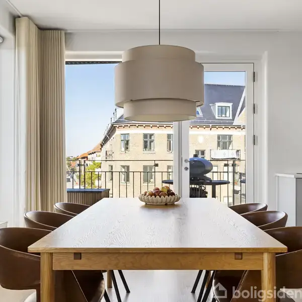 Et stort spisebord med otte læderstole står i et moderne spisestueområde. En frugtskål pryder bordet. Udenfor ses en balkon med en grill og en udsigt over hustage.