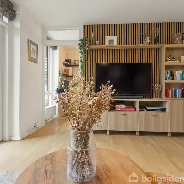 En vase med tørrede blomster står på et sofabord i en stue. Reolen med TV og bøger er bagved. Store vinduer til venstre slipper lys ind.