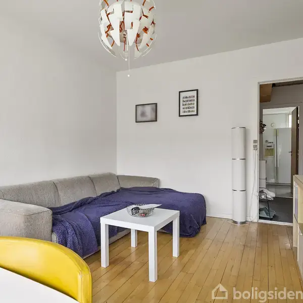 Grå sofa står i hjørnet, dækket af et blåt tæppe, ved siden af et hvidt sofabord. Gule stole omkranser et spisebord, og vægge prydes af kunst. Trægulv.