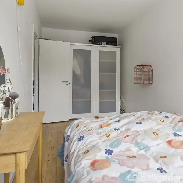 Soveværelse med enkeltseng dækket af farverigt sengetøj, placeret ved siden af et træbord med rund spejl og toiletartikler. I baggrunden ses en garderobe med glasdøre.