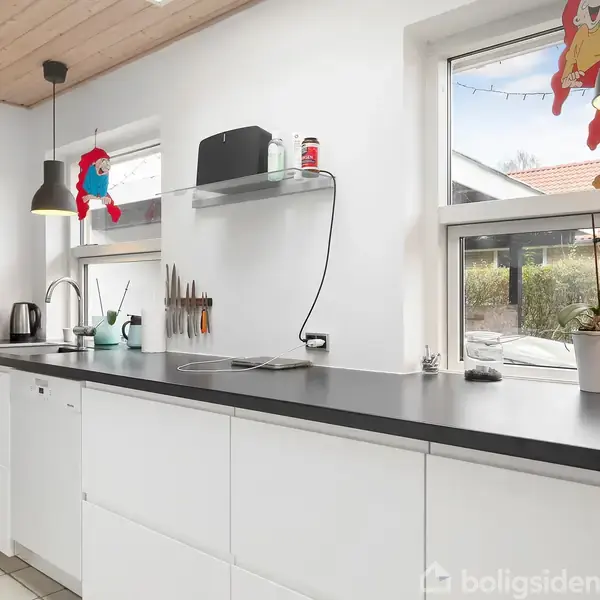 Køkkenbord med hvid vask, sæbedispenser og elkande; under hylden med krukke står knivholder. Lyse lamper hænger fra loftet. Store vinduer med udsigt til have omgivet af hække.