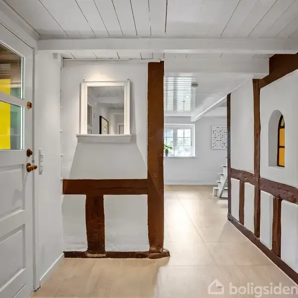 Dør med glaspartier åbner til en gang med hvide og brune bindingsværksvægge, klinkegulv og et spejl. Baggrunden viser et rum med et vindue og gulvplanter.