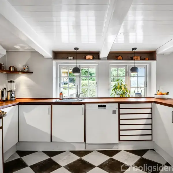 Køkken med hvide skabe og træbordplader. Sollys strømmer ind gennem vinduet over vasken, hvor der står en plante. Skakternet gulv og moderne lamper fuldender rummet.