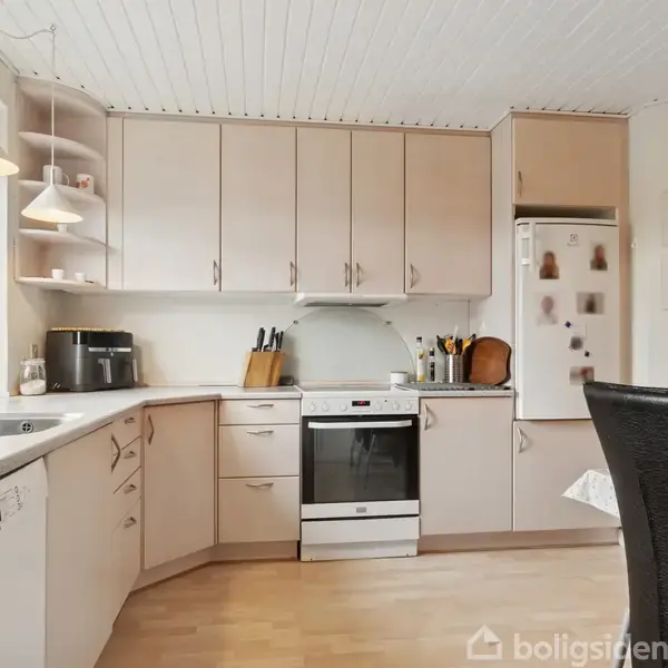 Køkkenet har lyse træskabe, en ovn midt i og køleskab til højre. En opvaskemaskine er på venstre side. To sorte stole står ved et bord til højre.