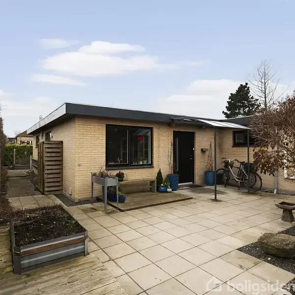 En enkeltstående murstensbygning med fladt tag står på en flisebelagt terrasse. Huset har store vinduer og omgivende hække samt cykler og krukker nær indgangen.