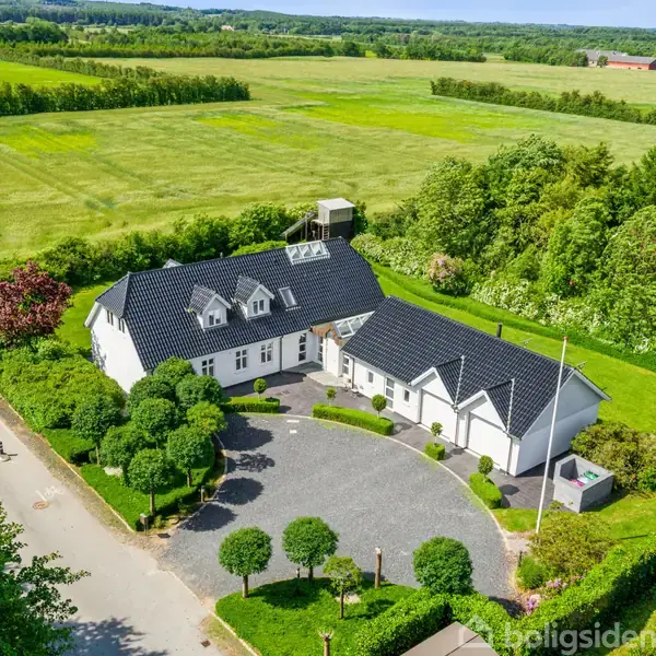 En hvid landejendom med sorte tage er omgivet af grønne marker. Den står ved en vej med tæt beplantning rundt om gårdspladsen, omgivet af velholdt buskads og træer.
