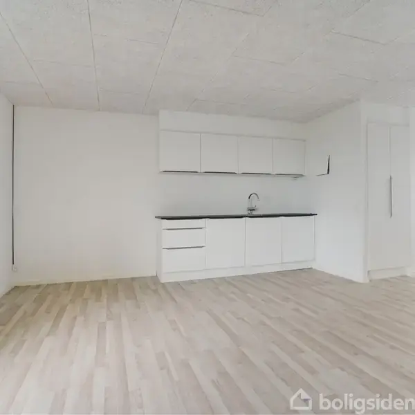 Et lyst køkken uden møbler har hvide skabe og en sort bordplade. Rummet har lyse trægulve og er åbent mod en tilstødende gang.