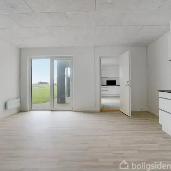 Et lyst rum med trægulv og hvide vægge har en åben dør til et køkken til højre. Glasdør fører ud til grønne marker udenfor.