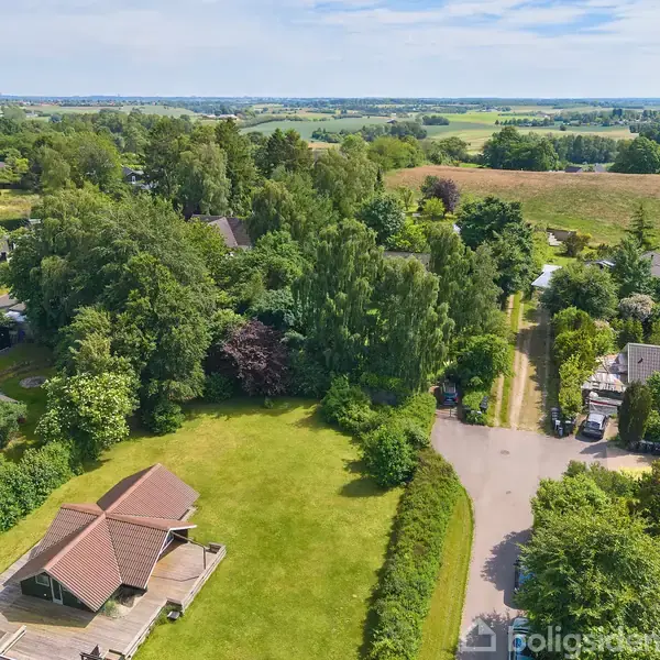 Hus med brunrøde tagsten står stille i en grøn have omkranset af træer. Omkring ligger flere huse og en vej i et frodigt landskab med skove og marker i baggrunden.