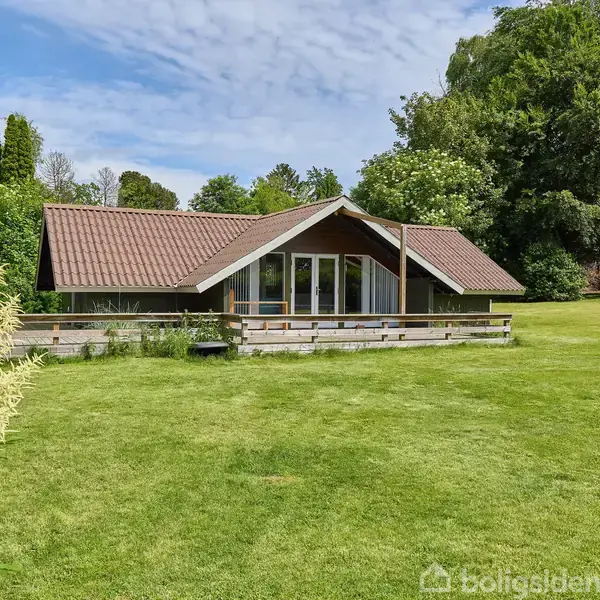 Et hus med træfacade og rødt tag står stille på en velplejet græsplæne, omgivet af tætte grønne træer under en klar blå himmel.