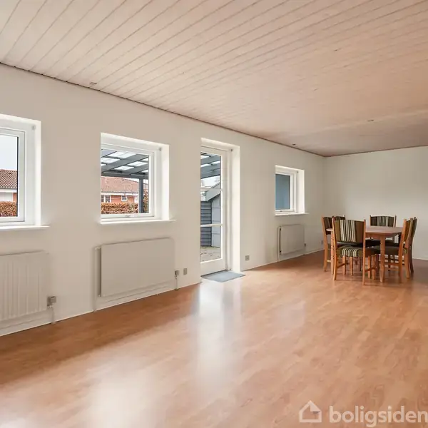 Stue med trægulv og hvidt loft, stillet op med et spisebord og stole. Rummet har fire vinduer og en dør, der fører ud til en overdækning.