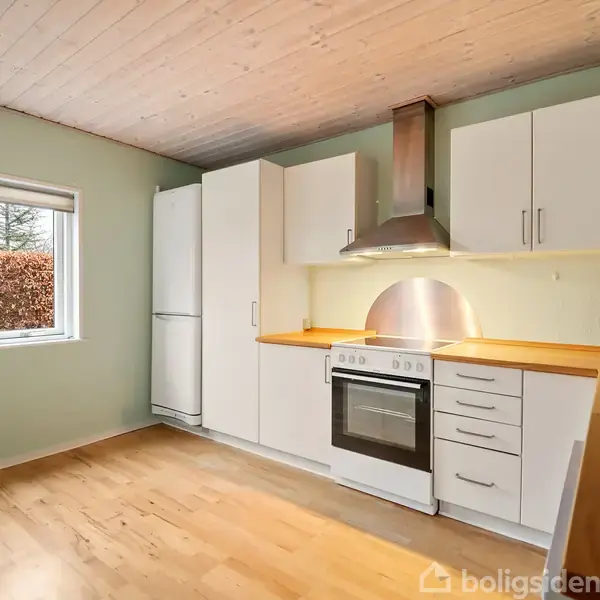 Køkken med komfur og emhætte indbygget i hvide skabe, placeret i et rum med lyst trægulv. Et stort vindue til venstre giver udsigt til en have med træer.