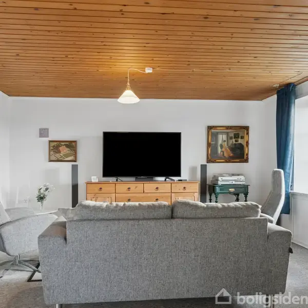 Stue med grå sofa og to stole står foran et tv på en trækonsol. Væggene har malerier, og et vindue med blå gardiner giver naturligt lys. Træloft og beige gulvtæppe.