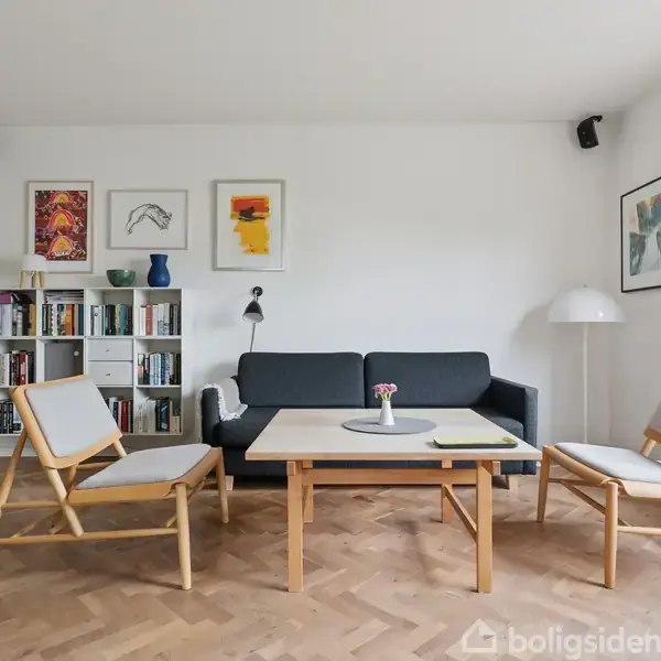 En stue med en sort sofa og to træstole omkranser et sofabord. Væghængt kunst og bogreol skaber en hyggelig atmosfære. Parketgulv og stor gulvlampe fuldender rummet.