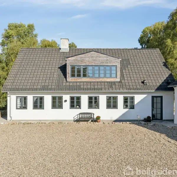 Et hvidt hus med gråt tag står på en grusbelagt gårdsplads. Et lille skur er til højre, og grønne træer omgiver ejendommen i baggrunden.