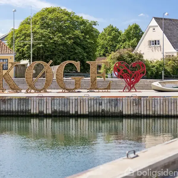 Store bogstaver staver "KØGE" med et hjerte som 'Ø'. De står ved en kaj med både foran flere huse og grønne træer i baggrunden.