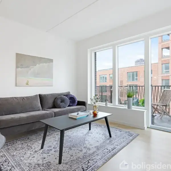 Gråt sofaarrangement med sofabord og tæppe foran store vinduer, der fører ud til en altan med udsigt til moderne bygninger. En gulvlampe og planter skaber en hyggelig atmosfære.