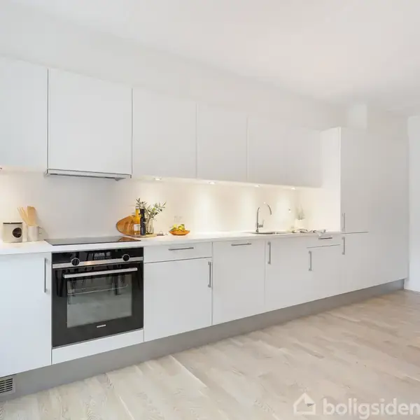 Et moderne køkken med hvide skabe og en ovn under kogepladen. Lige til venstre ses glas med tørvarer. Et rundt plantehjørneparadis er placeret til højre.