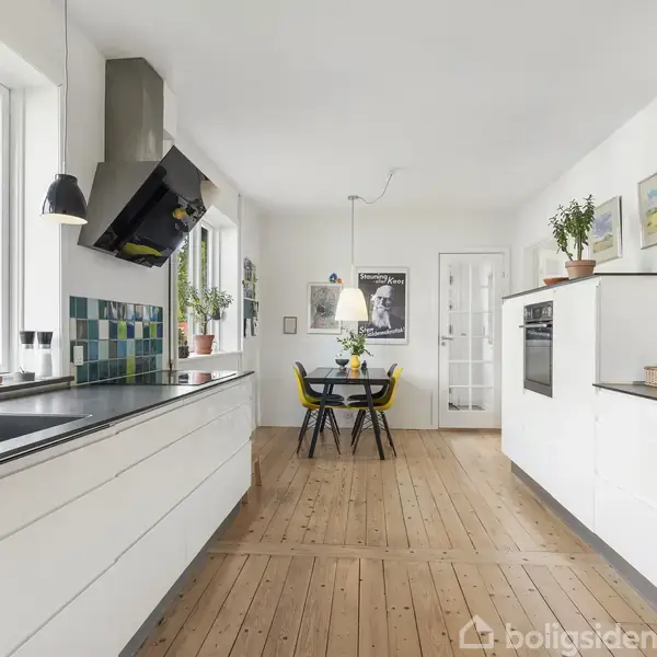 Køkken med hvide skabe og sort bordplade. Spisebord med fire stole er synlige for enden. Vinduer til venstre giver naturligt lys. Grønt udenfor og planter på bordpladen.