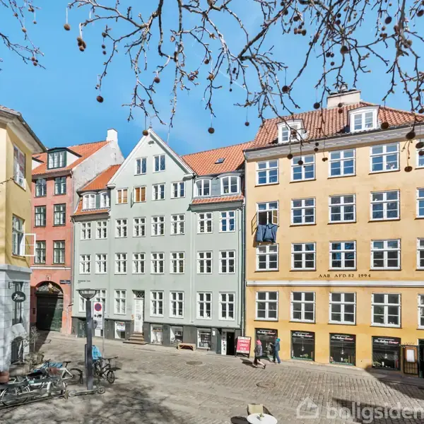 Farverige bygninger står oplyst af solen i en åben plads. Blade hænger fra en grænse af trægrene. Gaden indeholder cykler, og teksten "ANNO APRIL 1796" ses på den gule bygning.